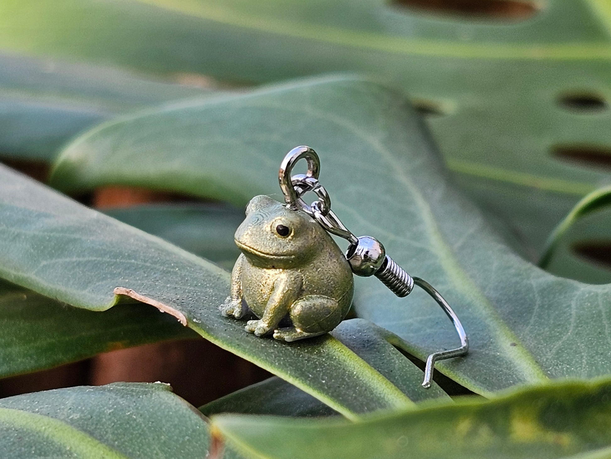 Tiny hand painted frog earring resting on a green leaf – Buck the Frog jewelry by Ghost Cactus Co